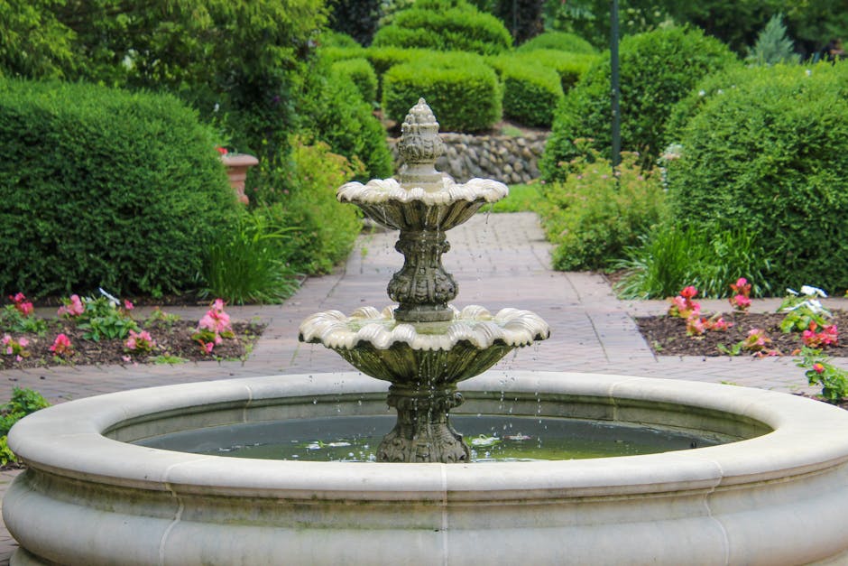 A decorative stone fountain surrounded by shrubs and flowers in a serene garden on a sunny summer day.